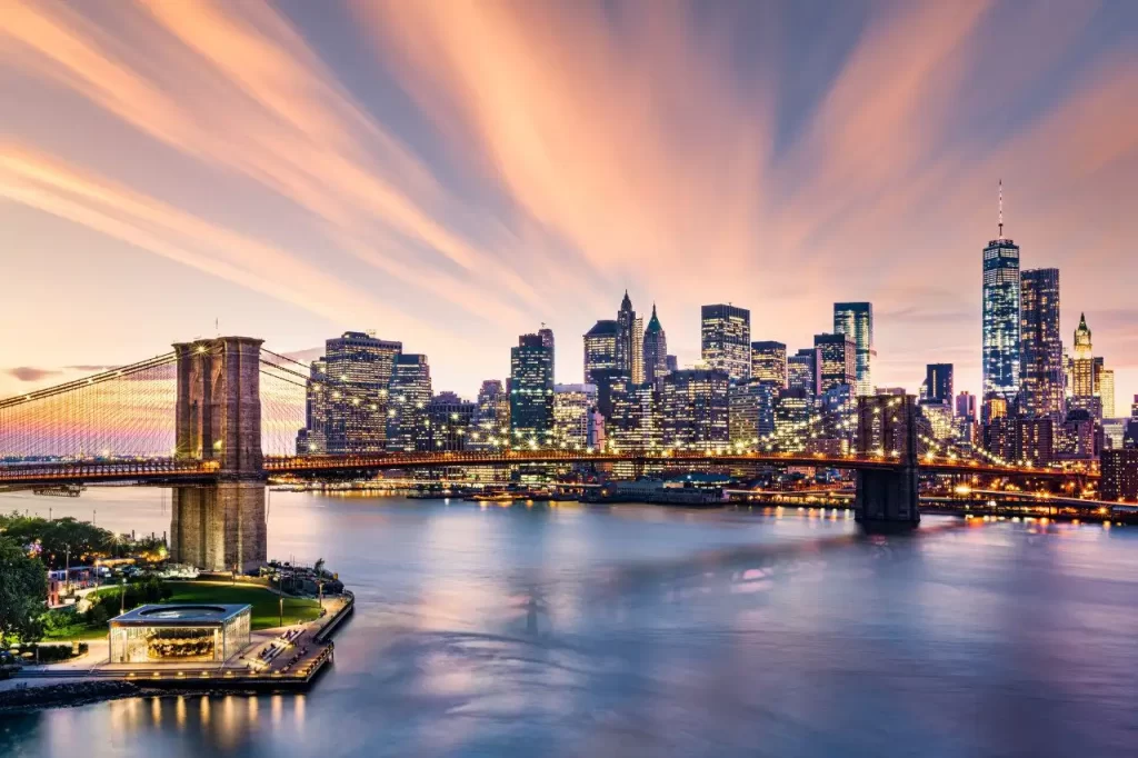 This is a scenic sunset view of the Brooklyn Bridge and Manhattan skyline in New York City, with reflections on the East River.