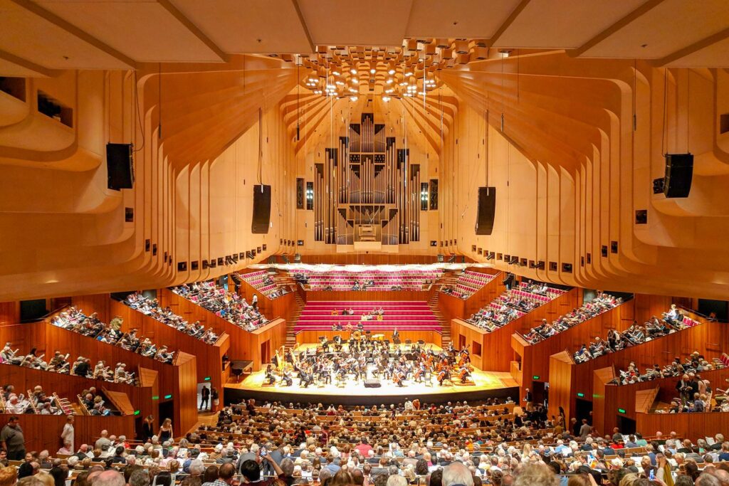 Interior view of the Sydney Opera House Concert Hall with a live orchestra performance and a seated audience enjoying the show"