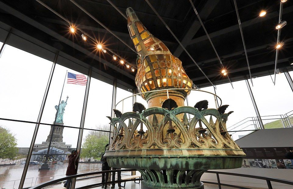 Original torch of the Statue of Liberty is displayed inside the Liberty Island Museum, with the statue and American flag visible through glass windows. Statue of Liberty Symbol of Freedom