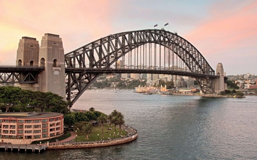 "Sydney Harbour Bridge spanning across the harbor at sunset with waterfront buildings, palm trees, and Luna Park in the background"