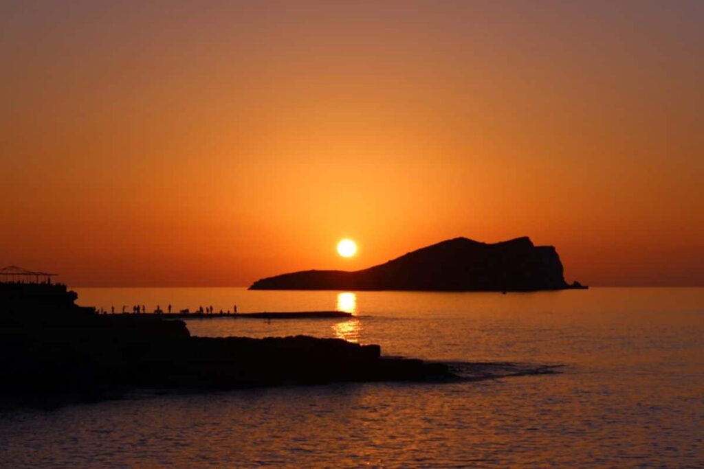 "Silhouetted island and beachgoers at sunset in Ibiza, Spain, with vibrant orange sky and calm Mediterranean waters reflecting the sun"