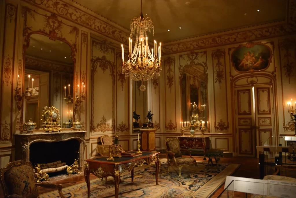 "Lavishly decorated royal palace room with ornate gold furniture, crystal chandelier, marble fireplace, and candlelit mirrors in classic European style"