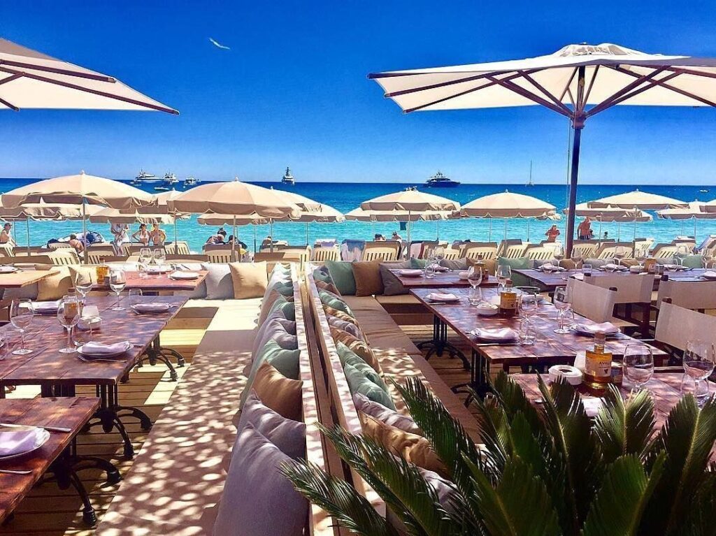 Luxury beachside restaurant with umbrellas, yachts, and turquoise waters on the French Riviera under a clear blue sky