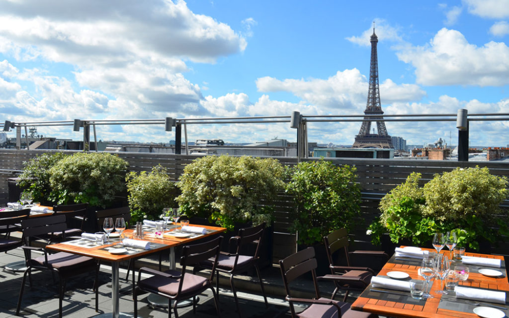 Rooftop restaurant in Paris with elegantly set tables and a scenic view of the Eiffel Tower under a partly cloudy blue sky
