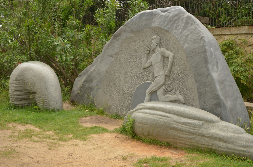 "Sculpture of a male runner emerging from a stone tunnel at Park Guell in Barcelona, Spain, surrounded by greenery and symbolic stonework"