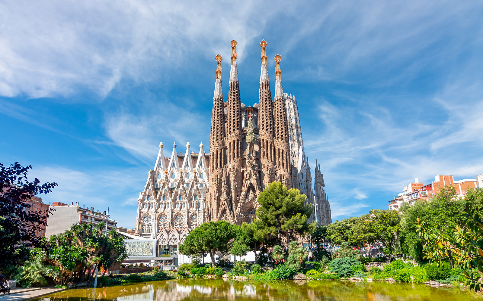 "Exterior view of the Sagrada Familia basilica in Barcelona, Spain, with its iconic spires and intricate facades reflected in a pond under a clear blue sky"