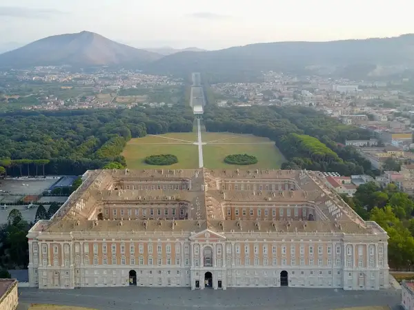 "Aerial view of the Royal Palace of Caserta in Italy, showcasing its grand symmetrical architecture, vast gardens, and distant mountains under a soft sunset sky"