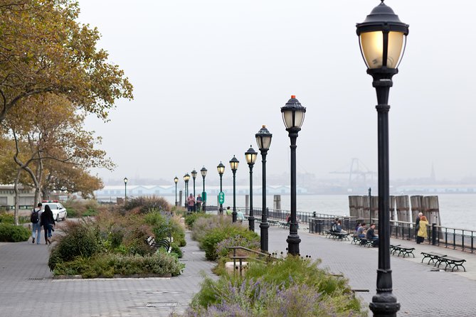 The Battery Park waterfront promenade in New York City offers a scenic view, complete with vintage lamp posts, lush greenery, and pedestrians strolling along the harbour. It's near Statue of Liberty Symbol of Freedom