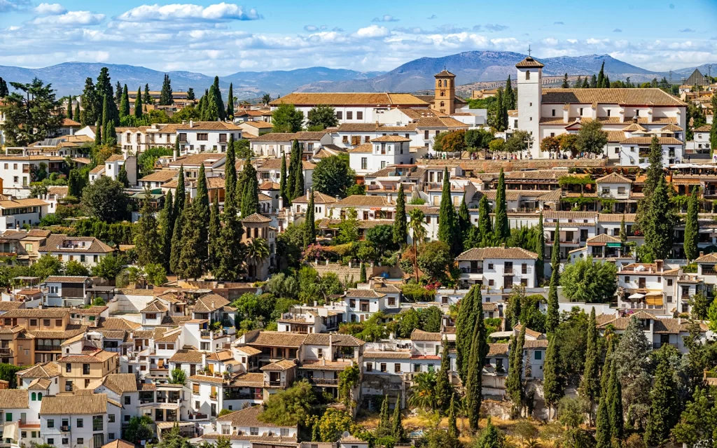 Alhambra Granada travel guide: "Panoramic view of the Albaicín district in Granada, Spain, with whitewashed houses, cypress trees, and historic churches set against the Sierra Nevada mountains"