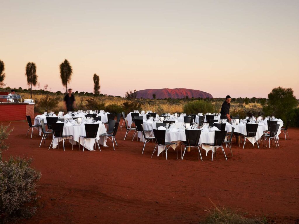 Outdoor fine dining setup on red desert sand with white tablecloths and Uluru (Ayers Rock) in the background at sunset in Australia's Red Centre
