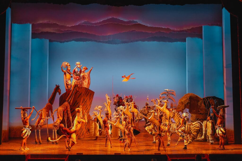 Broadway stage performance of The Lion King musical with vibrant costumes, animal puppets, and dramatic lighting during the iconic opening scene
