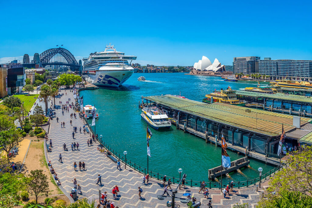 "A cruise ship docked at Circular Quay with Sydney Harbour Bridge, Opera House, and ferry terminals in view on a bright sunny day."