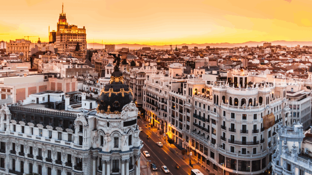 "Aerial view of Gran Vía and the Metropolis Building in Madrid, Spain, during a vibrant sunset with golden sky and city lights"