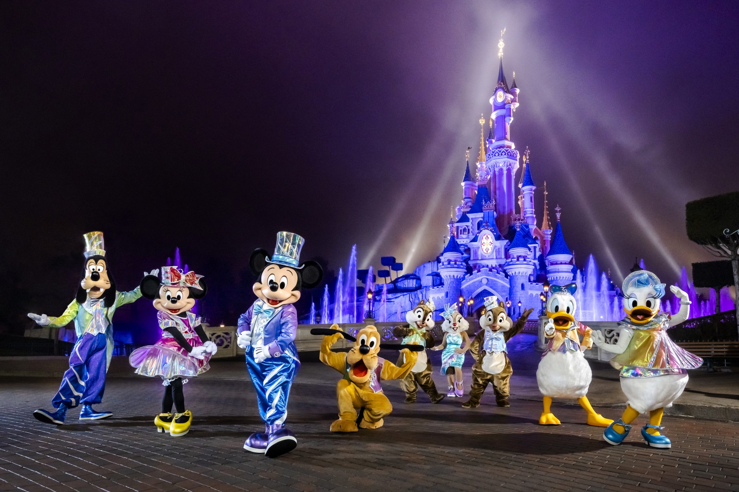 Disneyland Paris travel guide: "Mickey Mouse, Minnie Mouse, Donald Duck, Goofy, Pluto, and Disney characters in sparkling costumes posing in front of the illuminated Sleeping Beauty Castle at Disneyland Paris during a nighttime show."
