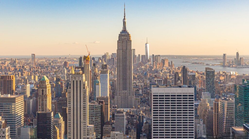 The Empire State Building towers above the Manhattan skyline during the day, with One World Trade Centre and the Hudson River in the background.