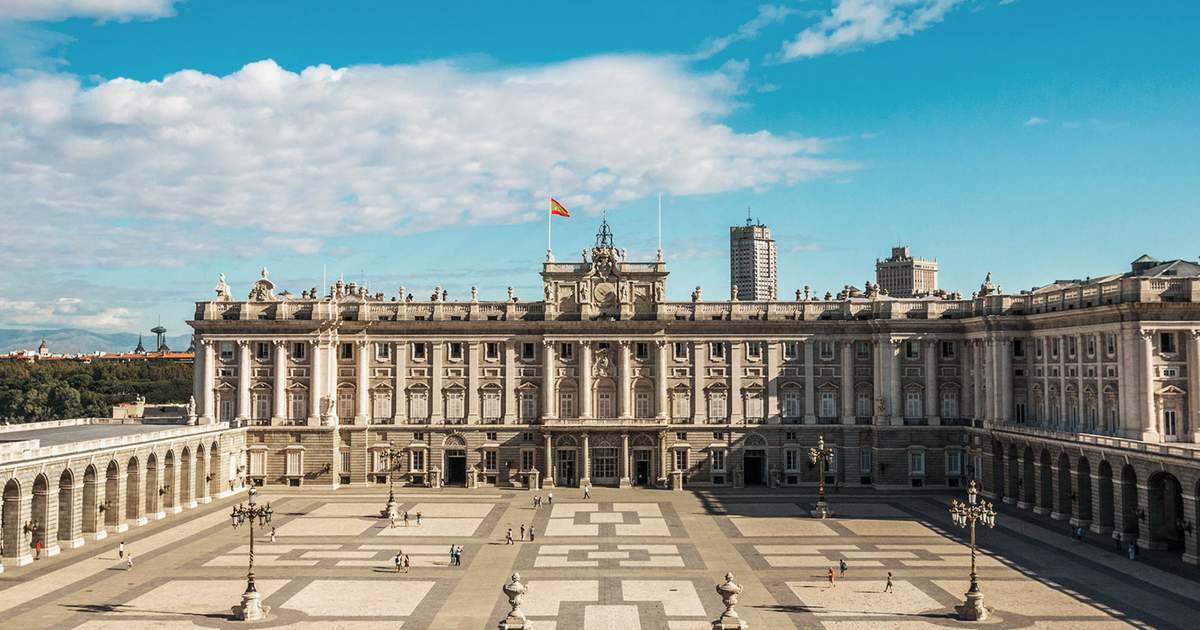 "Panoramic view of the Royal Palace of Madrid with the Spanish flag flying above, showing the grand courtyard and neoclassical architecture under a bright blue sky"