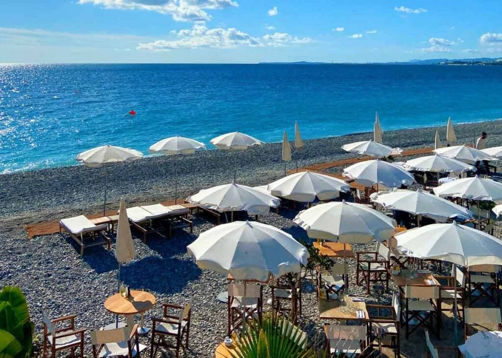 French Riviera travel guide: Pebble beach in Nice, France with white sun umbrellas, lounge chairs, and clear blue Mediterranean Sea under a sunny sky