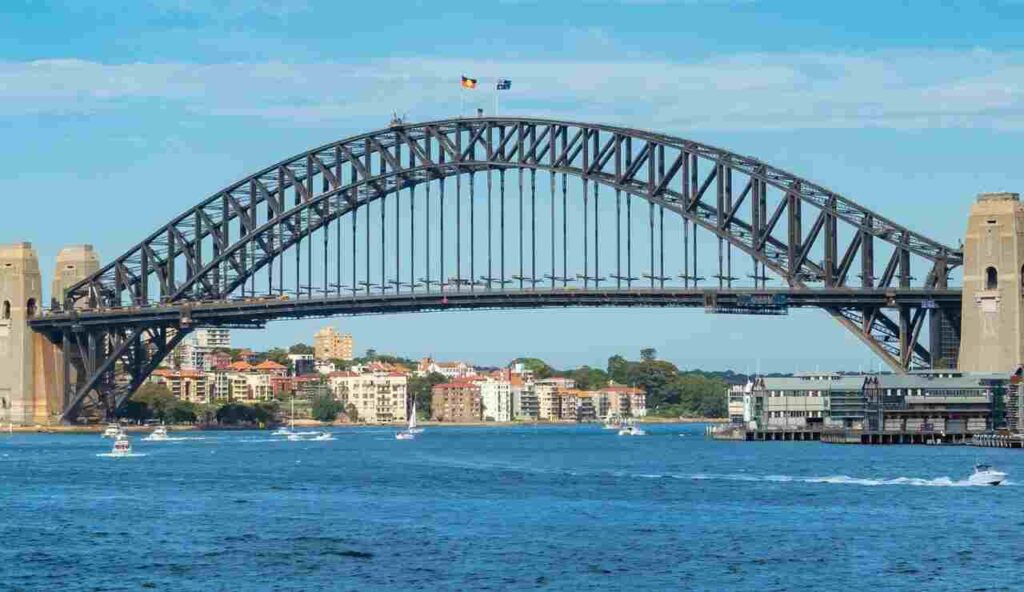 "Sydney Harbour Bridge spanning across the harbor with boats sailing below and city skyline in the background on a sunny day"