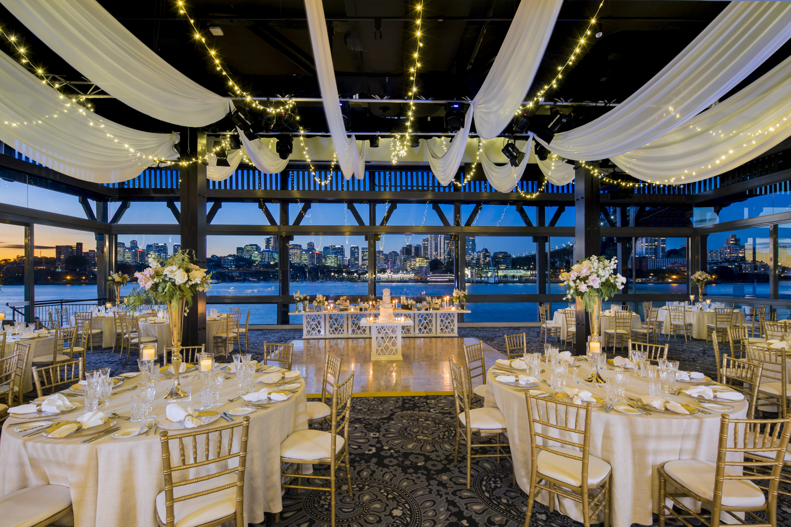 Luxurious waterfront wedding reception venue in Sydney with decorated tables, draped ceilings, and city skyline views at sunset