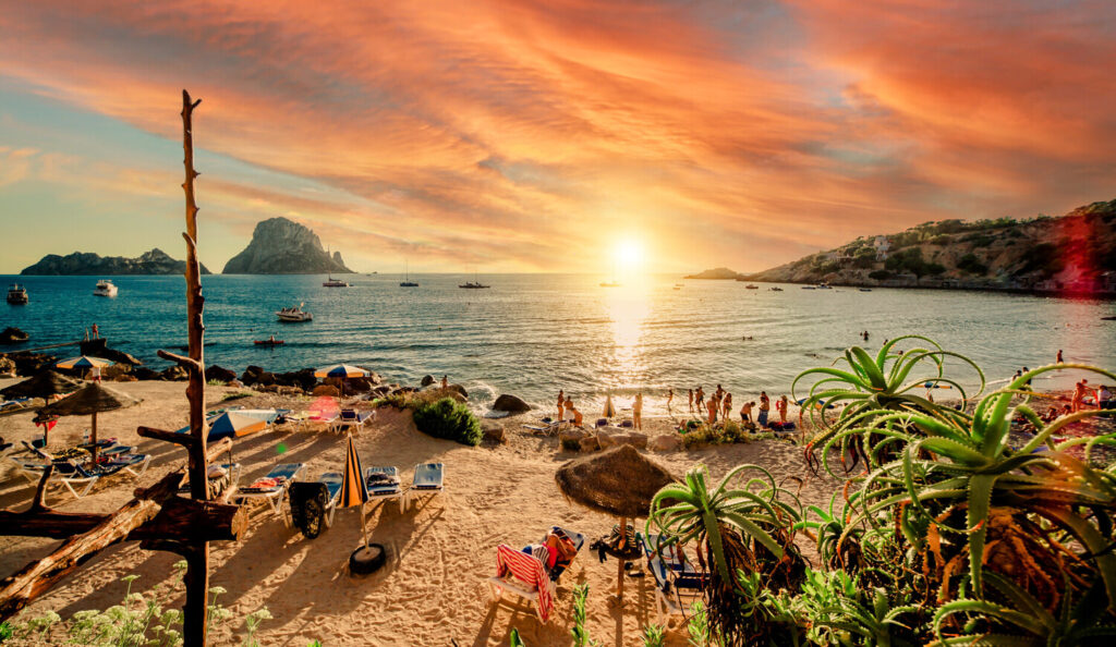 "Golden sunset over Cala d’Hort beach in Ibiza, Spain, with people enjoying the sand, sun loungers, and scenic views of Es Vedra island in the distance"