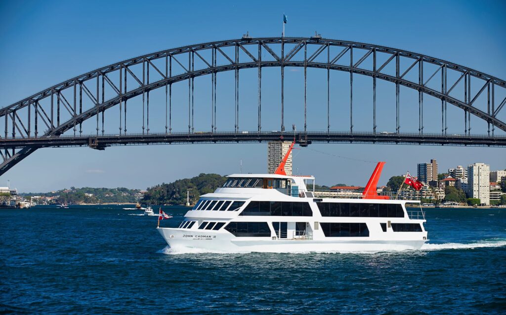Sydney Harbour Bridge climb or cruise: Sightseeing cruise boat John Cadman II sailing under the Sydney Harbour Bridge with city buildings in the background on a clear blue day"