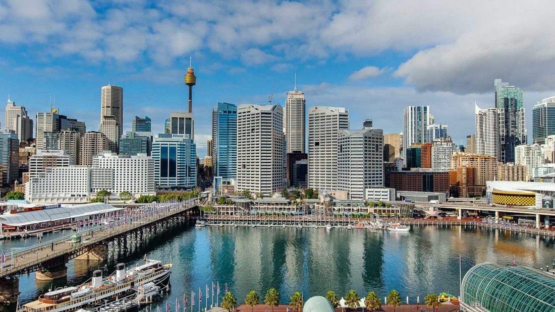 Panoramic view of Darling Harbour in Sydney with waterfront buildings, boats, and city skyline under a partly cloudy sky