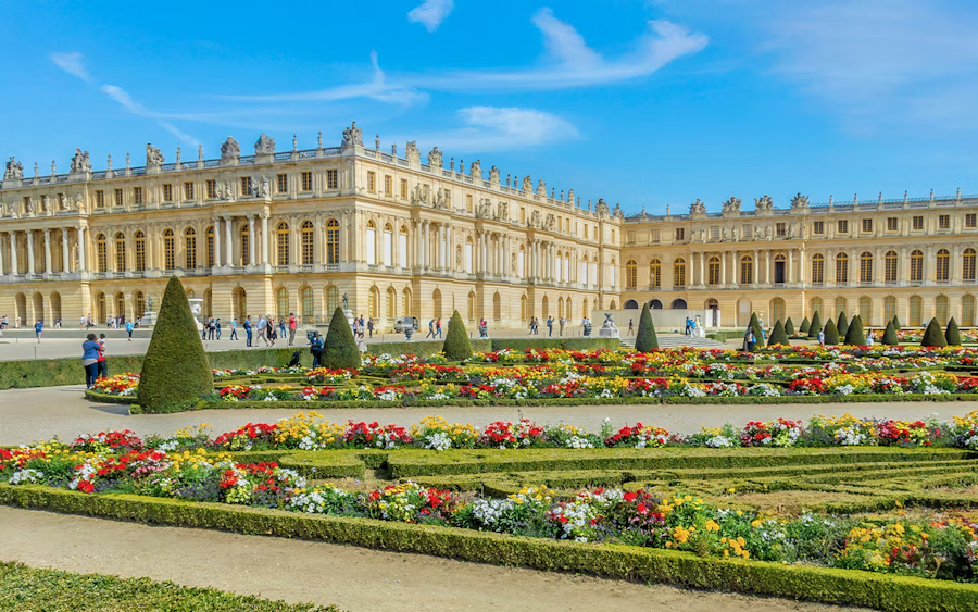 View of the Palace of Versailles in France with vibrant formal gardens, symmetrical flowerbeds, and historic baroque architecture under a clear blue sky