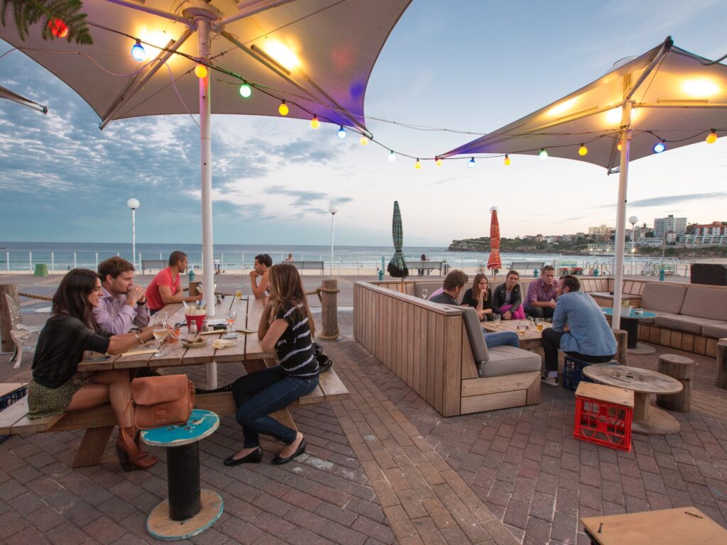 People dining and relaxing at a beachside café with ocean views at Bondi Beach, Sydney, under colorful lights during sunset