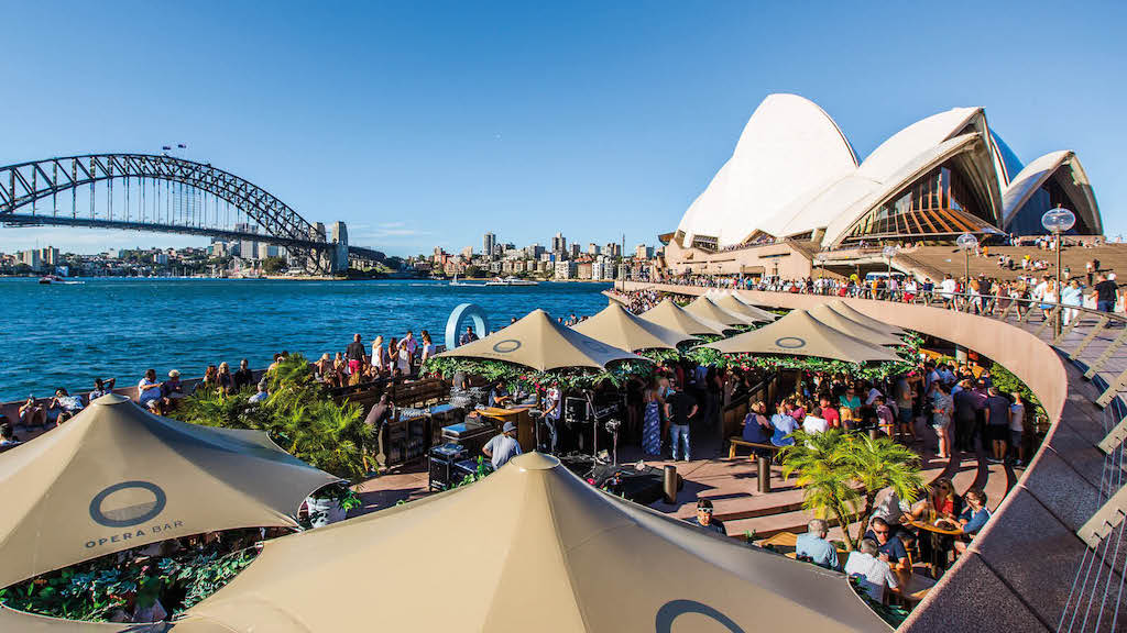 Crowds enjoying food and drinks at Opera Bar near the Sydney Opera House with views of Sydney Harbour Bridge and the waterfront skyline on a sunny day