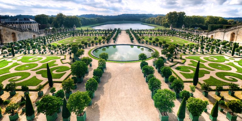 Chateau de Versailles travel guide: Aerial view of the symmetrical Orangerie Gardens at the Palace of Versailles, France, featuring formal landscaping, fountains, and tree-lined paths