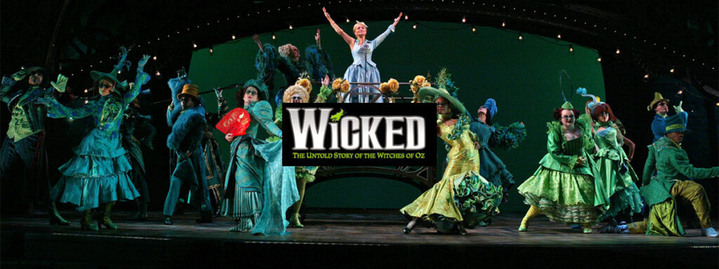 Broadway cast of Wicked performing on stage in vibrant green costumes with Glinda, the lead character, in the spotlight at centre stage.