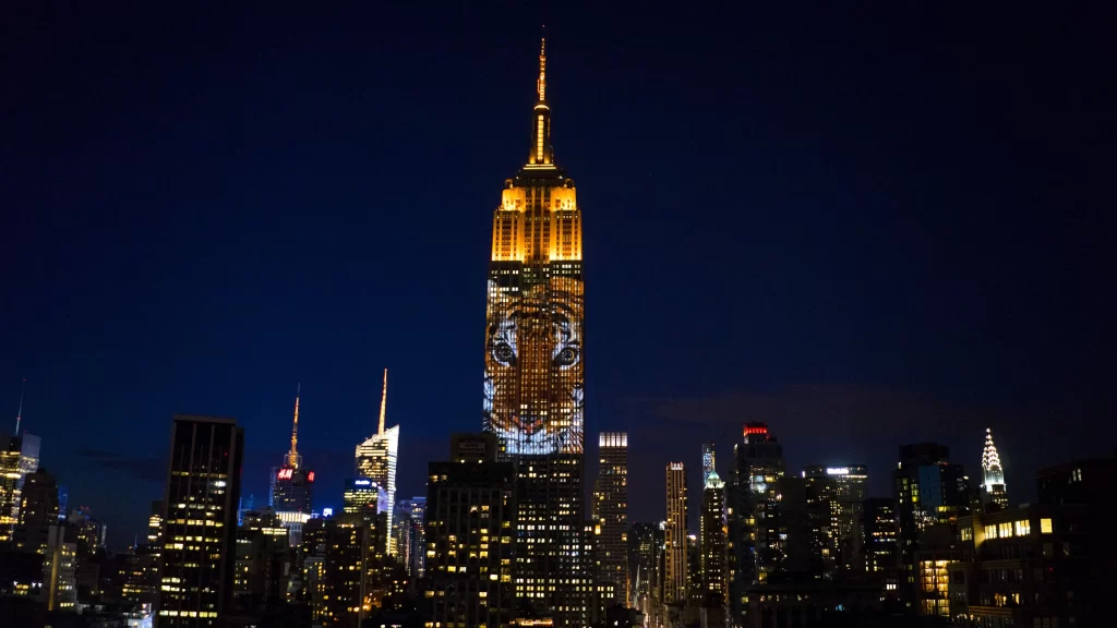 The Empire State Building lit up at night with a glowing tiger face projection during a wildlife awareness event, surrounded by New York City skyline.