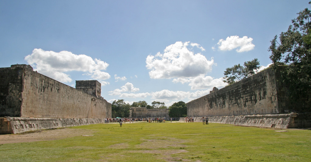 Chichen Itza travel guide: "The Great Ball Court at Chichen Itza, Mexico, with massive stone walls and grassy playing field under a blue sky with clouds"