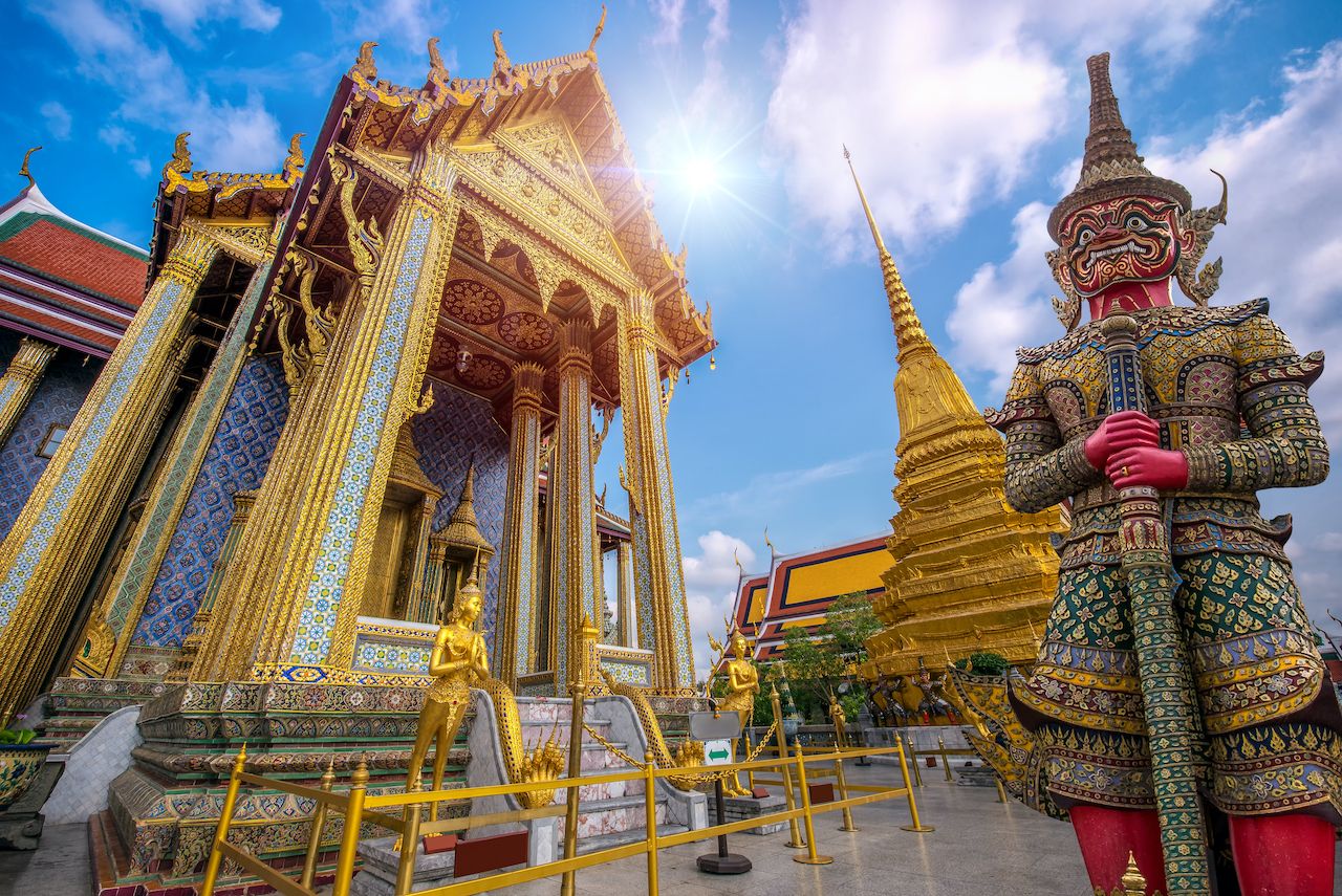 Grand Palace in Bangkok: "Colourful guardian statue and golden temple structures at Wat Phra Kaew in the Grand Palace, Bangkok, Thailand, under a bright blue sky."