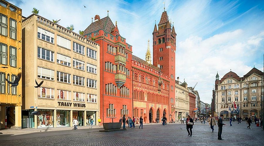 Cologne Cathedral travel guide: "People walking through the historic Marktplatz in Basel, Switzerland, with the red Basel Town Hall (Rathaus) and surrounding buildings under a blue sky"