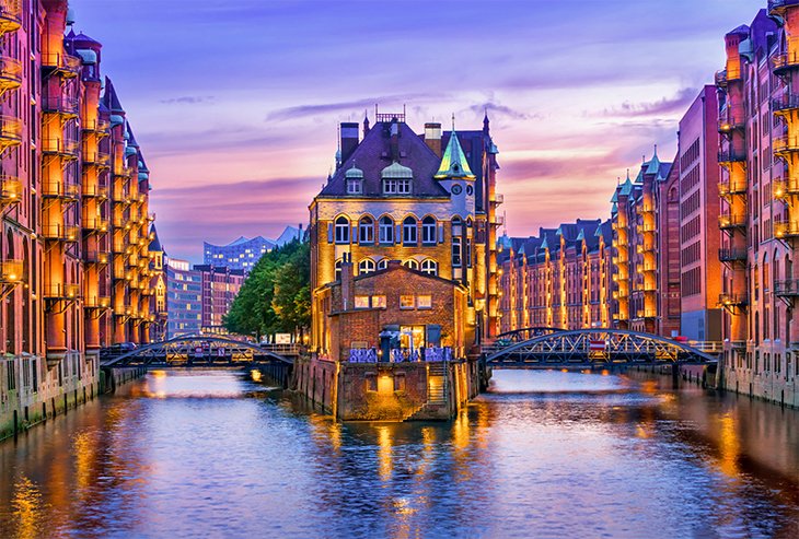 "Evening view of the illuminated Speicherstadt warehouse district in Hamburg, Germany, with historic brick buildings and canal reflections under a vibrant purple sunset sky"