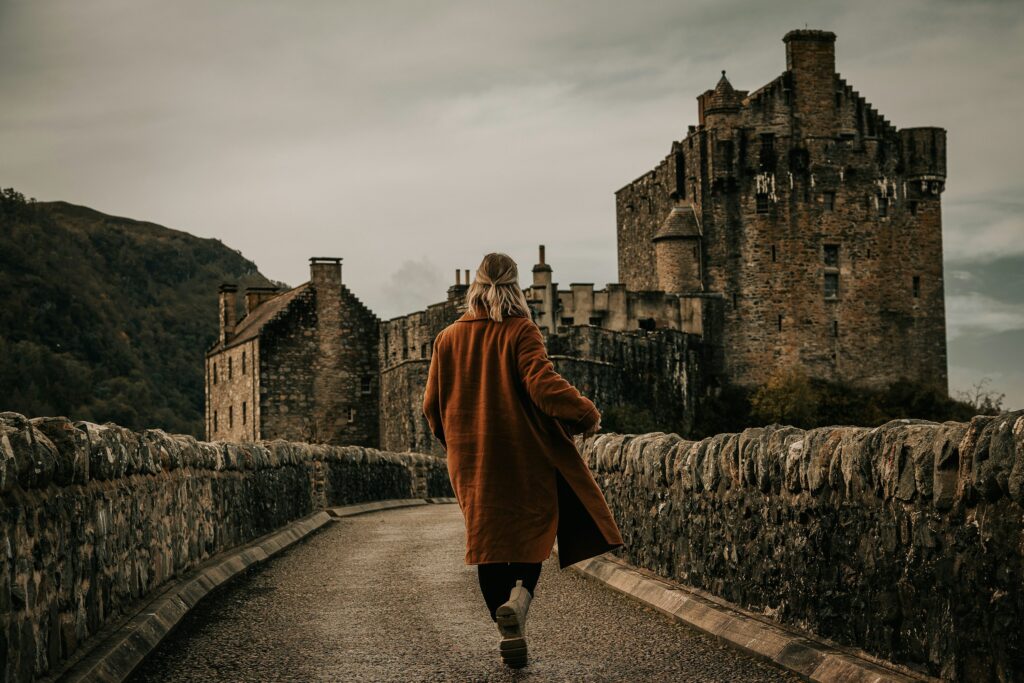 "Woman in a long brown coat walking toward a medieval stone castle on a cloudy day, crossing a historic stone bridge"