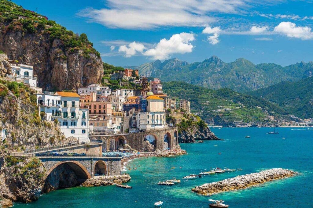 "Beautiful coastal view of Amalfi town in Italy with colorful houses built on cliffs, turquoise Mediterranean waters, boats in the harbor, and green mountain backdrop"