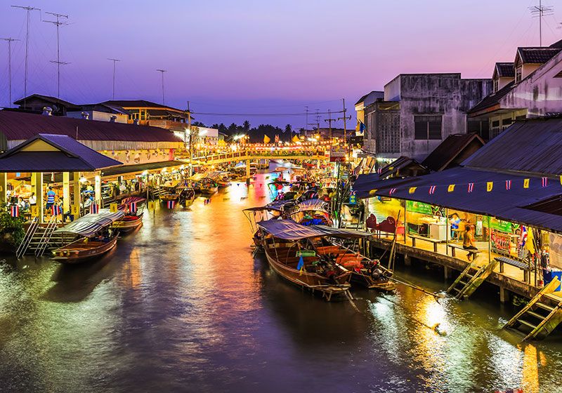 "Evening view of Amphawa Floating Market in Thailand with colorful boats, riverside shops, and glowing lights reflecting on the water at sunset"