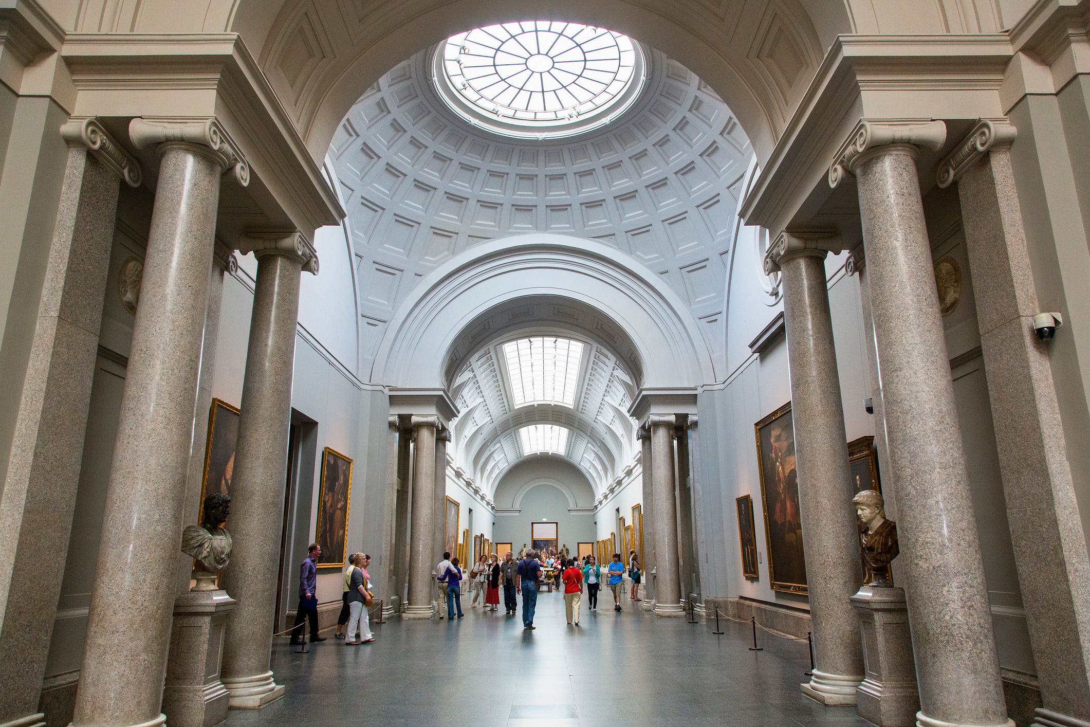 "Visitors walking through the grand arched gallery hall of the Prado Museum in Madrid, Spain, featuring classical columns, skylights, and European paintings on display"