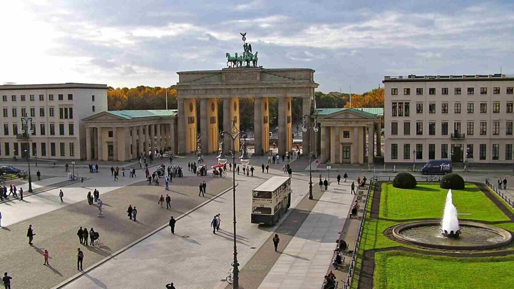 "Brandenburg Gate in Berlin, Germany, with tourists walking around Pariser Platz and a Berlin City Tour bus passing in front on a cloudy afternoon"