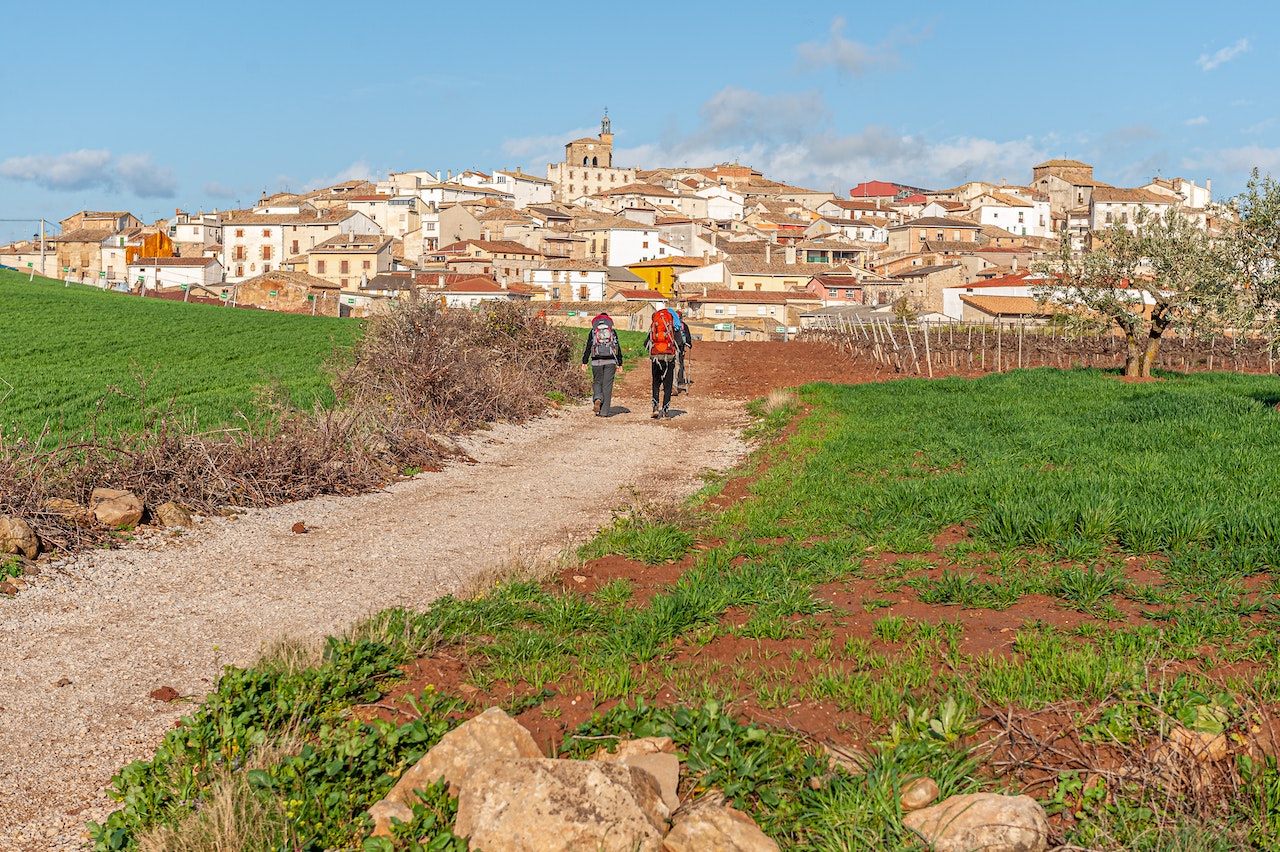 Camino de Santiago spiritual journey: "Pilgrims walking along the Camino de Santiago trail toward a traditional Spanish village under a clear blue sky surrounded by green countryside fields"