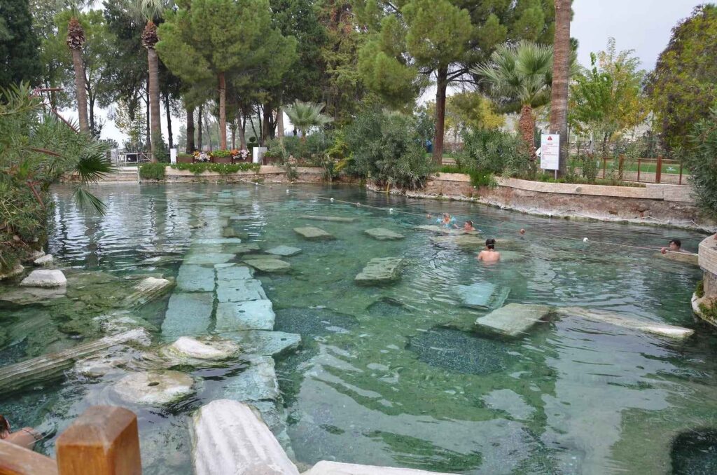"Tourists swimming in Cleopatra’s Pool, the ancient thermal spring in Pamukkale, Turkey, surrounded by lush greenery and submerged Roman columns in crystal-clear water"