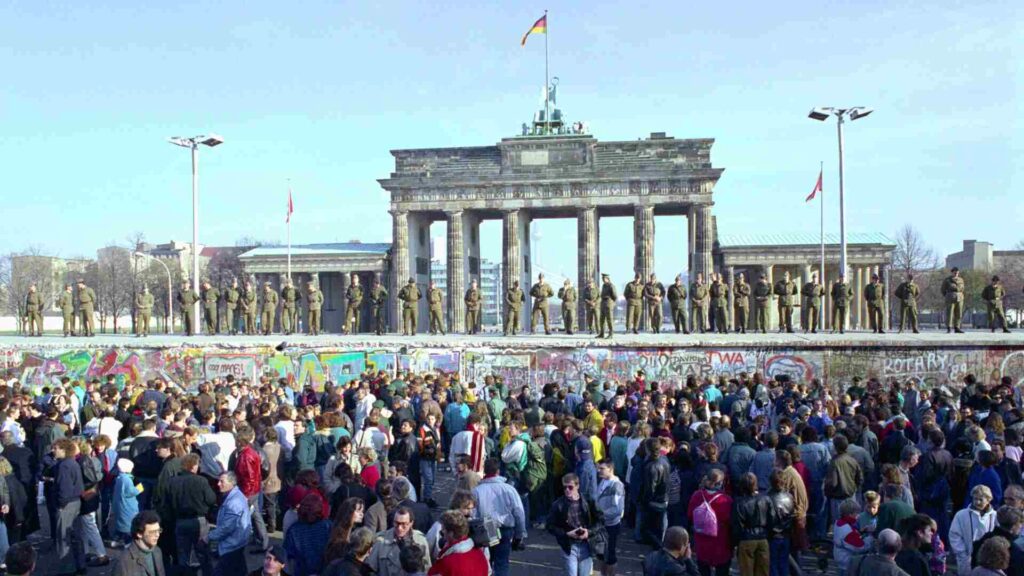 Brandenburg Gate Berlin: "Historic photo of the Berlin Wall in front of the Brandenburg Gate with East German border guards standing on top and crowds celebrating the fall of the wall in 1989"