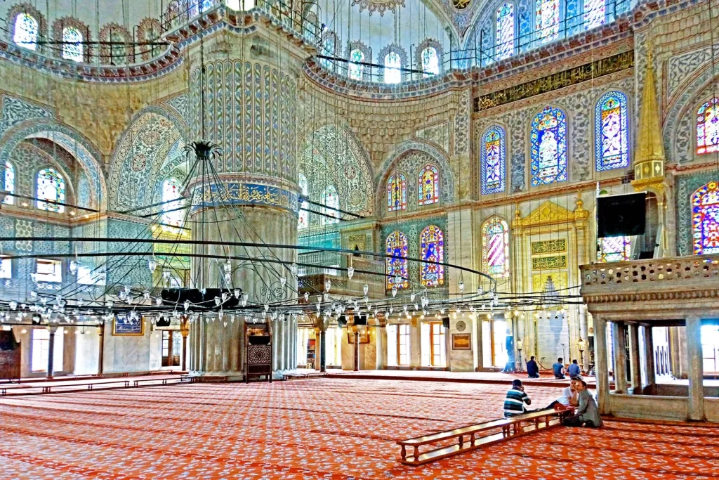 "Interior of the Blue Mosque in Istanbul, Turkey, showcasing intricate blue İznik tiles, stained glass windows, and worshippers inside the grand prayer hall"