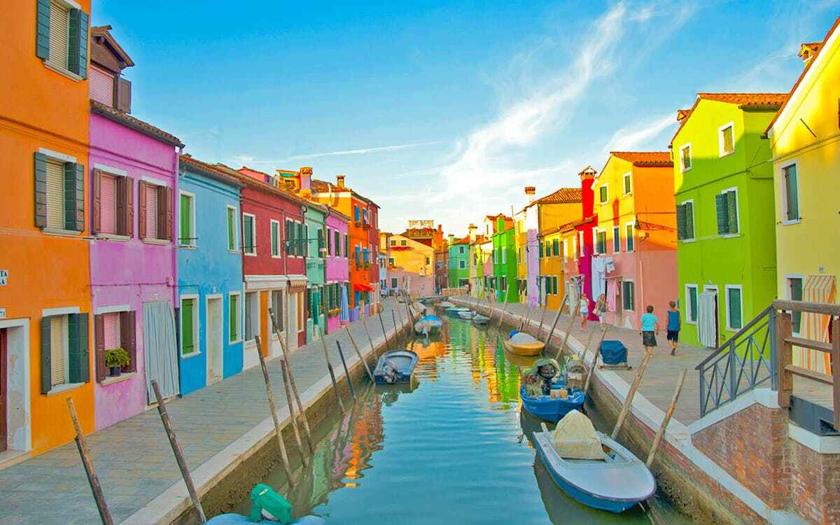 Venice canals travel guide: "Colorful houses along a quiet canal on Burano Island in Venice, Italy, with small boats reflecting in the water under a bright blue sky"