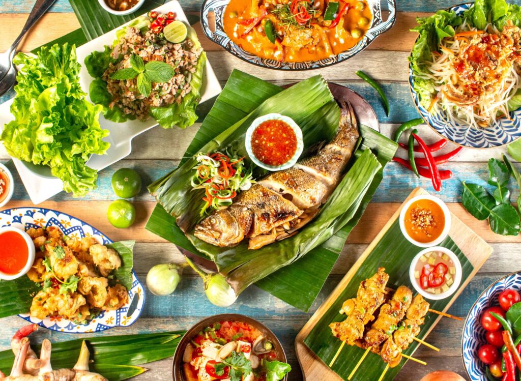 "Traditional Thai cuisine with grilled fish, satay skewers, spicy curries, fresh herbs, and sauces served on banana leaves"