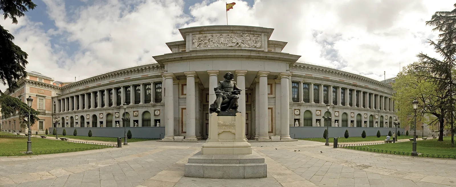 "Front view of the Prado Museum in Madrid, Spain, featuring the Velázquez statue, neoclassical architecture, and Spanish flag under a cloudy sky"
