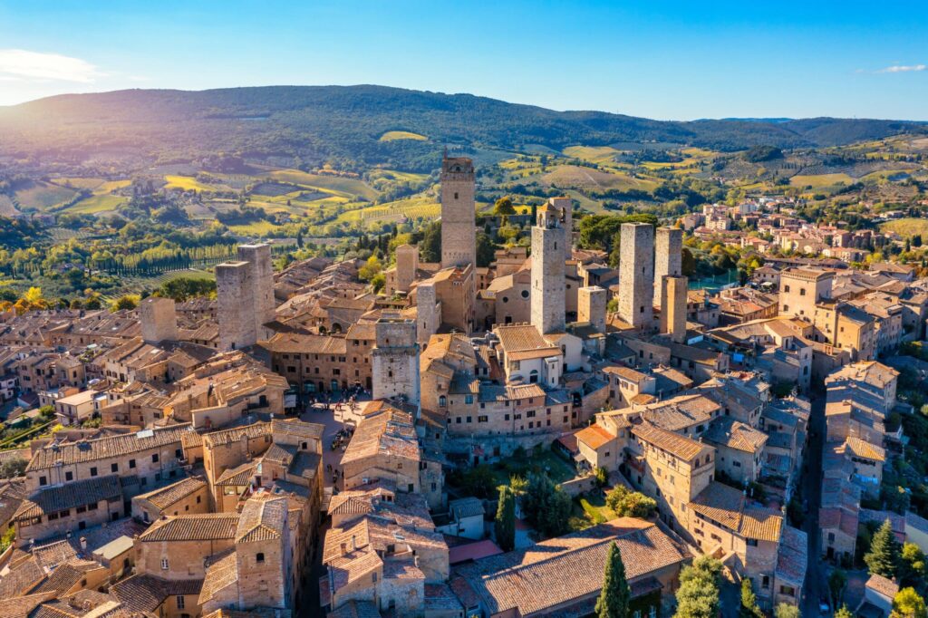 Tuscany vineyards travel guide: "Aerial view of San Gimignano in Tuscany, Italy, featuring medieval stone towers, terracotta rooftops, and rolling green hills under a clear blue sky"