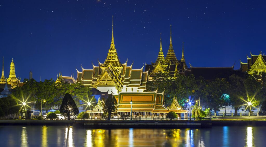 "Grand Palace in Bangkok, Thailand illuminated at night with golden rooftops reflecting on the Chao Phraya River under a starry sky"